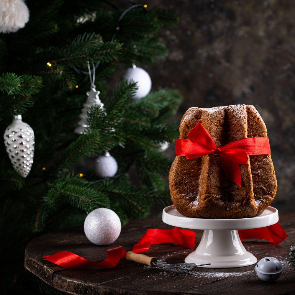 Traditional Italian pandoro Christmas cake tied with a red ribbon, displayed beside a decorated tree.